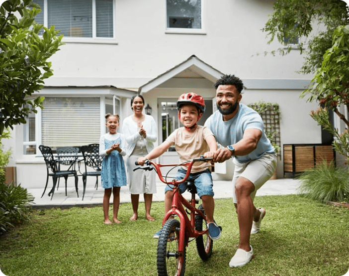 A man helps a young boy ride a bicycle on grass, while two women and a girl stand nearby in front of a house, smiling and clapping.