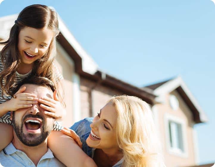 A man carries a smiling girl on his shoulders while she covers his eyes; a woman beside them laughs. They enjoy a sunny day outside their new home, thanks to the help of long-distance movers.