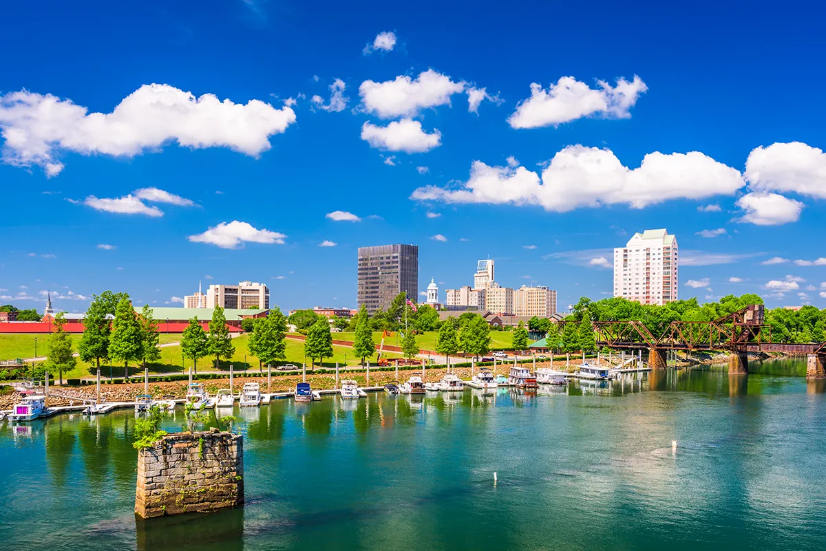 A waterfront cityscape in Savannah, with boats docked along the river, green trees lining the shore, and buildings under a blue sky—this scenic view is a favorite spot for movers in Georgia to unwind after a busy day.