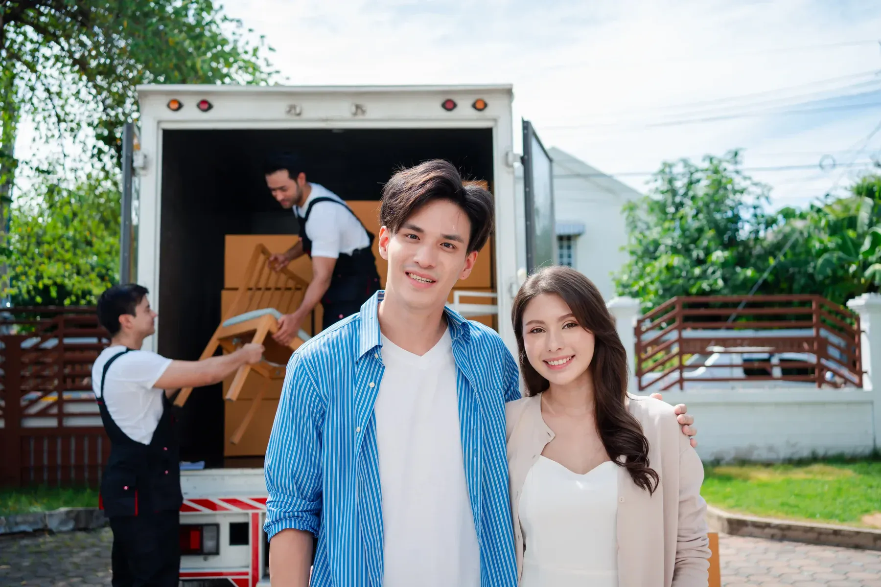 A smiling couple stands in front of a moving truck as vetted moving companies load cardboard boxes, ensuring you protect your move in a friendly residential neighborhood.
