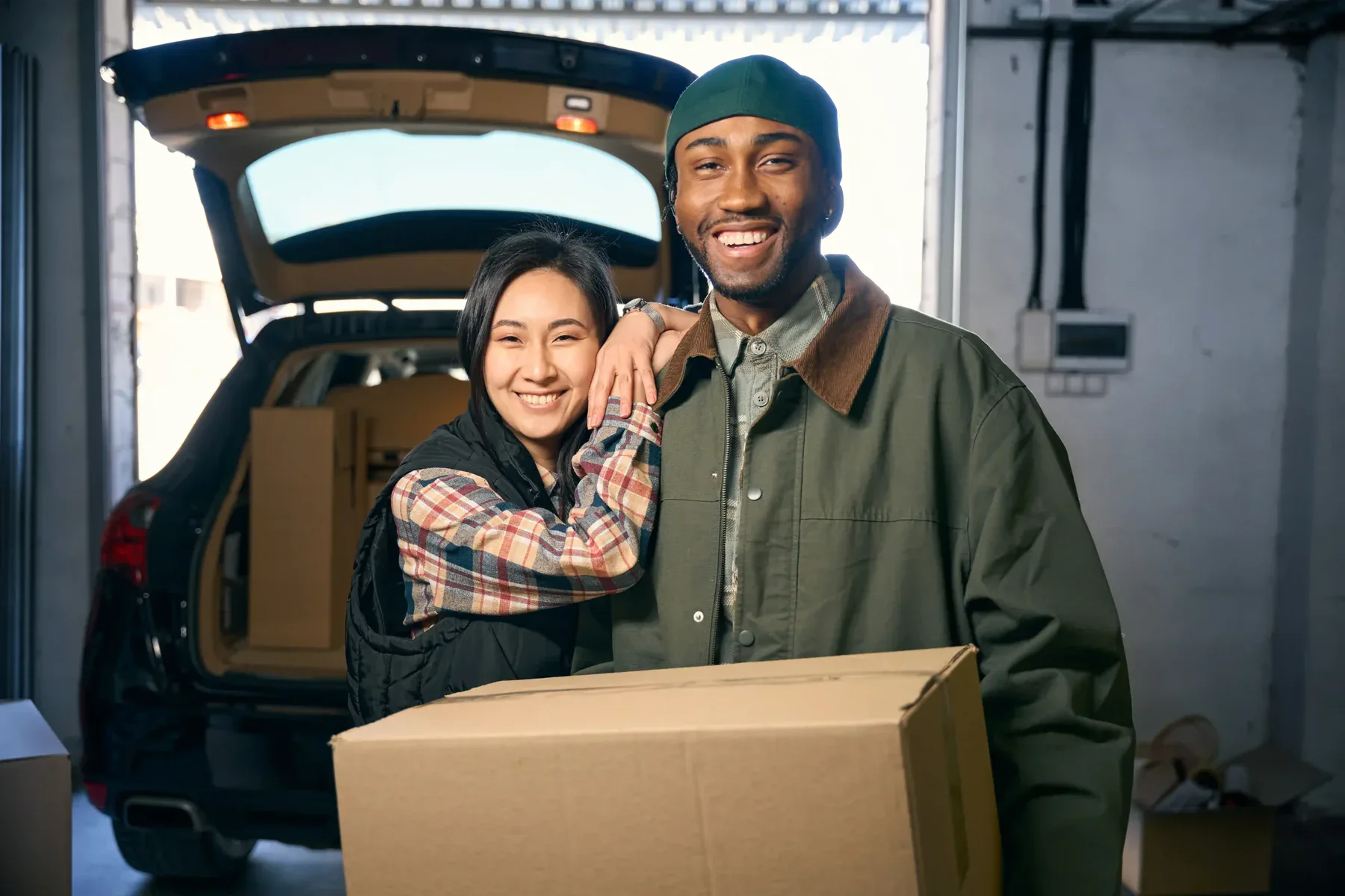 Two people smiling, standing in front of an open car trunk filled with boxes. One person is holding a cardboard box—they look ready to move with money-saving movers in a garage or storage area.